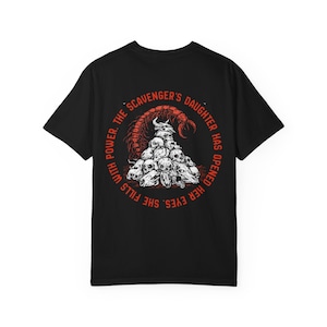 May include: A black short-sleeve t-shirt featuring a circular graphic on the back. The design displays a pile of skulls topped with a horned skull and a large scorpion, surrounded by the text: THE SCAVENGER'S DAUGHTER HAS OPENED HER EYES. SHE FILLS WITH POWER.