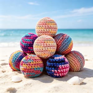 May include: A pyramid of seven colorful, crocheted balls on a sandy beach with seashells. The balls are various colors, including pink, blue, yellow, and orange. The ocean and sky are visible in the background.