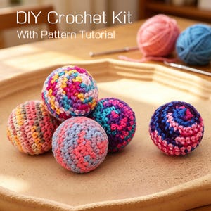 May include: A DIY crochet kit with a pattern tutorial. The image shows several colorful crochet balls in shades of pink, blue, purple, and orange. The balls are arranged on a light brown surface, with yarn and crochet hooks in the background.