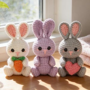 Op de afbeelding: Drie handgemaakte gehaakte konijnen in wit, lavendel en grijs. Het witte konijn houdt een wortel vast, het lavendel konijn heeft een eenvoudig ontwerp en het grijze konijn houdt een roze hart vast. Elk konijn heeft roze binnenoren en zwarte knoopogen.