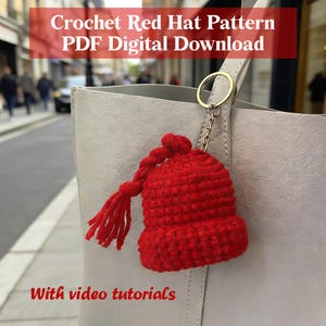 May include: A small, bright red crocheted hat charm with a tassel, hanging from a silver keychain attached to a light gray handbag. The text "Crochet Red Hat Pattern PDF Digital Download" and "With video tutorials" are visible.