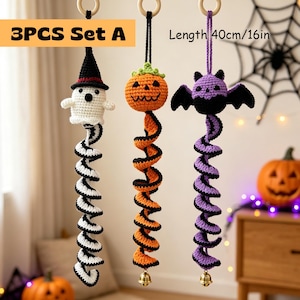 May include: A 3-piece set of hanging crochet decorations, each 40cm long. The set includes a white ghost with a black hat, an orange pumpkin, and a purple bat. Each figure features a long, spiraling tail and a small gold bell at the bottom.