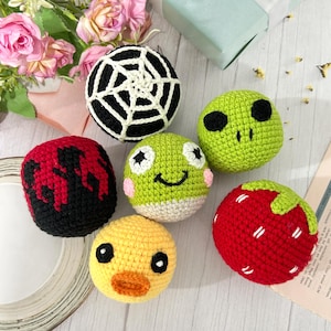 May include: Six round crocheted yarn balls featuring various designs: a black and white spider web, a green alien face, a red and black flame pattern, a green frog face with pink cheeks, a yellow duck face, and a red strawberry with green leaves.
