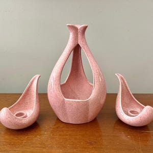 May include: A set of three pink ceramic decorative pieces. The set includes a tall, open-design vase and two matching candle holders. The pieces have a speckled texture and a mid-century modern aesthetic.