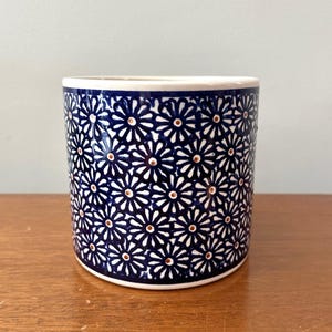 May include: A cylindrical ceramic container with a white rim and base. The exterior is covered in a repeating pattern of white daisy-like flowers with brown centers on a dark blue background. The container sits on a wooden surface.