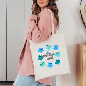 May include: White canvas tote bag with a blue and yellow design. The bag features the words "Hospice Aide" surrounded by blue flowers with words like "friendly", "loyal", "helpful", "empathetic", "smart", "kind", and "caring" written on the petals.