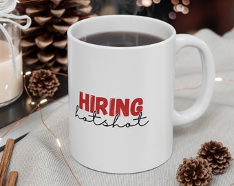 May include: White ceramic coffee mug with a black handle. The mug has the text "HIRING hotshot" printed on it in red and black lettering. The mug is sitting on a white fabric surface with a string of lights.