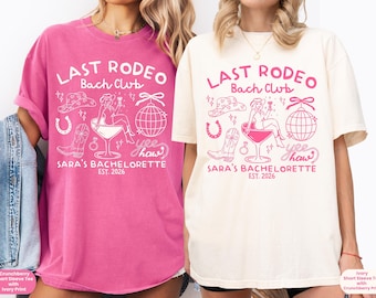 May include: Two short-sleeve t-shirts, one pink and one ivory, feature a white and pink graphic design. The design includes the text "LAST RODEO Bach Club" and "Sara's Bachelorette EST. 2026" with cowboy-themed illustrations. The shirts are a comfortable fit.