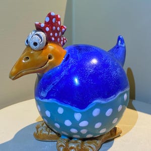 Könnte beinhalten: Eine skurrile Keramik-Hühnerfigur. Das Huhn hat einen blauen Körper mit weißen Punkten, einen orangefarbenen Schnabel und einen roten Kamm mit weißen Punkten. Die Figur sitzt auf braunen Füßen.