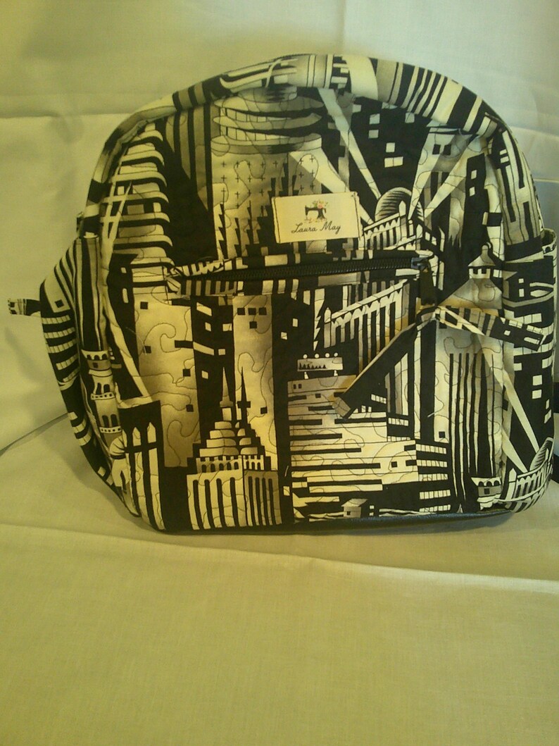 Peut inclure: Un sac &agrave; dos noir et blanc avec un motif de paysage urbain. Le sac a une poche avant zipp&eacute;e et une petite &eacute;tiquette avec le texte "Laura May" et un logo de machine &agrave; coudre. Le sac &agrave; dos est en tissu matelass&eacute;.