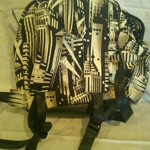 Peut inclure: Sac &agrave; dos noir et blanc avec un motif de paysage urbain. Le sac a des bretelles noires et une fermeture &eacute;clair. Le motif repr&eacute;sente des b&acirc;timents et des &eacute;l&eacute;ments architecturaux.