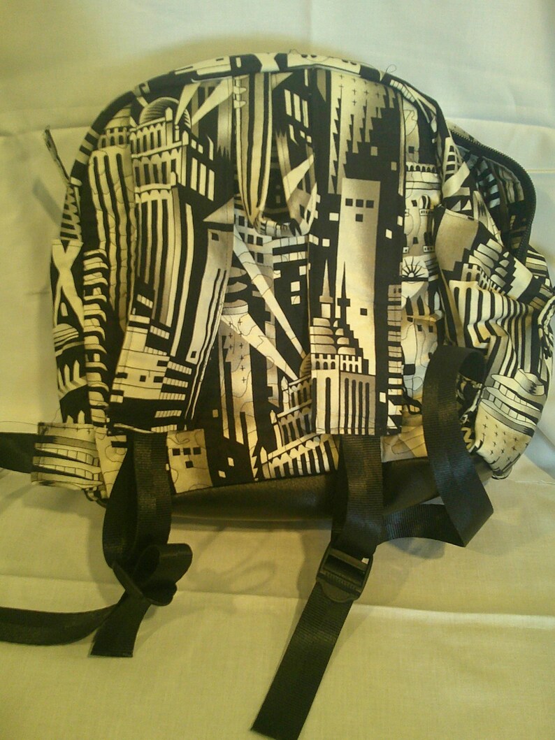 Peut inclure: Sac &agrave; dos noir et blanc avec un motif de paysage urbain. Le sac a des bretelles noires et une fermeture &eacute;clair. Le motif repr&eacute;sente des b&acirc;timents et des &eacute;l&eacute;ments architecturaux.