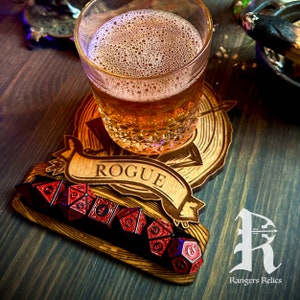 May include: A wooden coaster with a  "Rogue" banner and an arrow design. A glass of amber liquid sits on the coaster. Six red and black dice are arranged on the coaster.