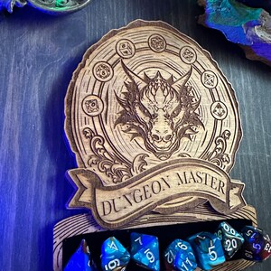 May include: Wooden Dungeon Master dice holder with a dragon design and a banner that reads "Dungeon Master". The holder has a slot for six dice, and there are six blue dice in the slot.