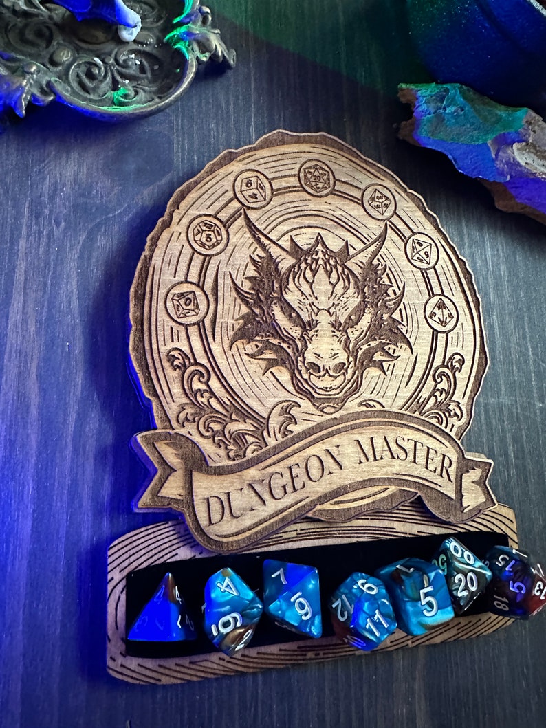 May include: Wooden Dungeon Master dice holder with a dragon design and a banner that reads "Dungeon Master". The holder has a slot for six dice, and there are six blue dice in the slot.