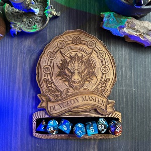 May include: A wooden dice tower with a dragon design and the text "Dungeon Master". The tower has a slot for holding dice, and there are seven blue and white dice in the slot.