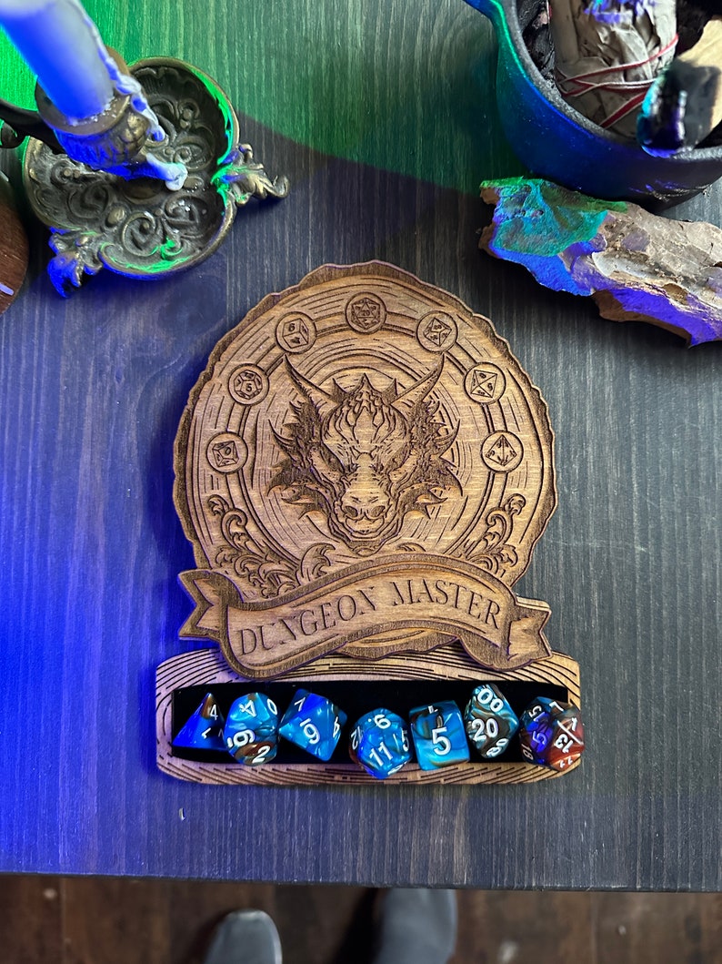 May include: A wooden dice tower with a dragon design and the text "Dungeon Master". The tower has a slot for holding dice, and there are seven blue and white dice in the slot.