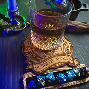 May include: A wooden dice tray with the words "Dungeon Master" engraved on it. The tray has a slot for holding dice and a small glass of amber liquid is sitting on top of the tray. There are several blue and black dice in the slot.