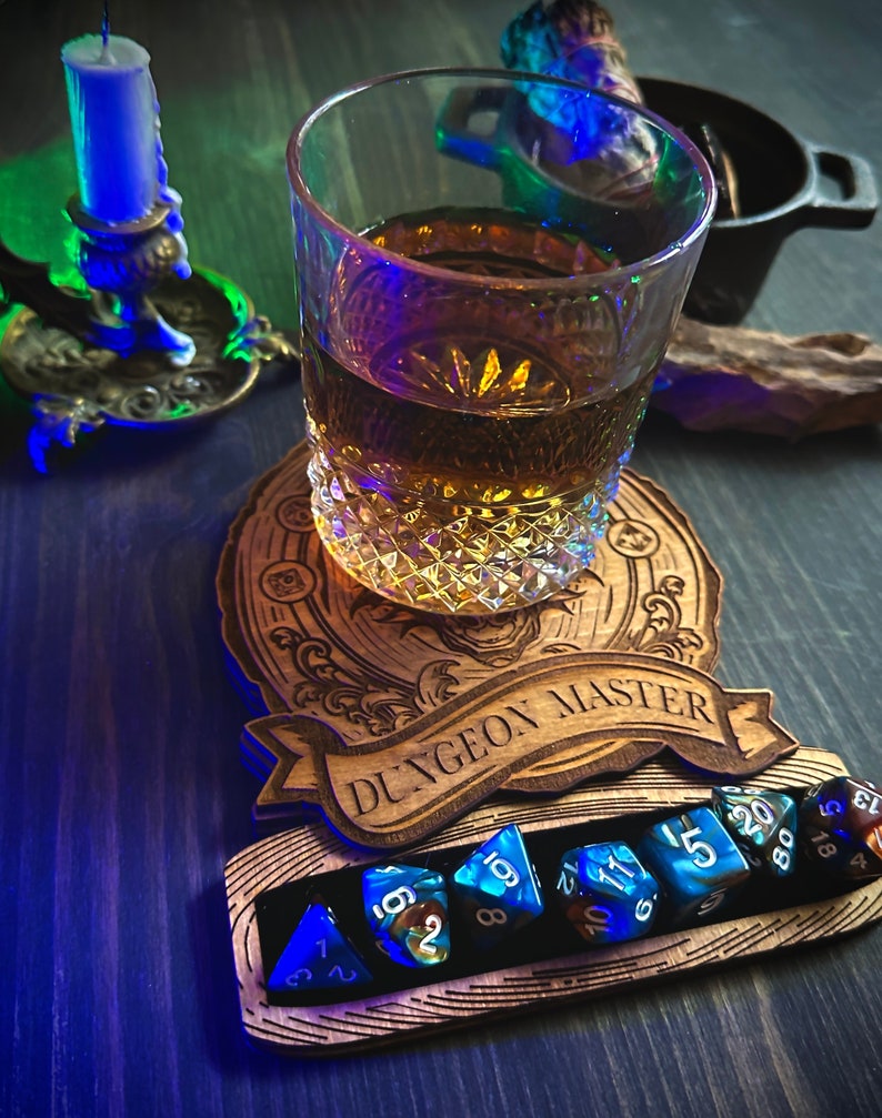 May include: A wooden dice tray with the words "Dungeon Master" engraved on it. The tray has a slot for holding dice and a small glass of amber liquid is sitting on top of the tray. There are several blue and black dice in the slot.