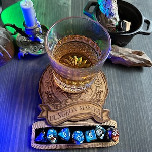 May include: A wooden coaster with the words "Dungeon Master" engraved on it. A glass of amber liquid sits on the coaster. Seven blue and brown dice are in a groove on the coaster.