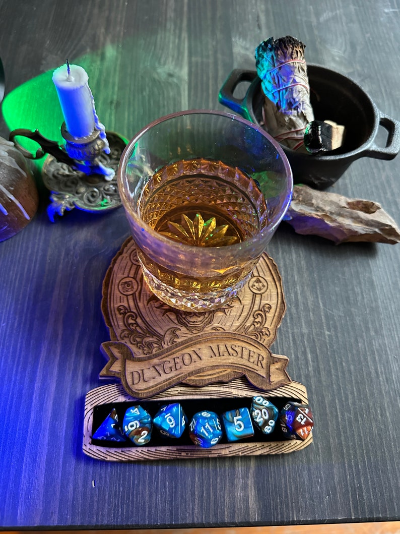 May include: A wooden coaster with the words "Dungeon Master" engraved on it. A glass of amber liquid sits on the coaster. Seven blue and brown dice are in a groove on the coaster.