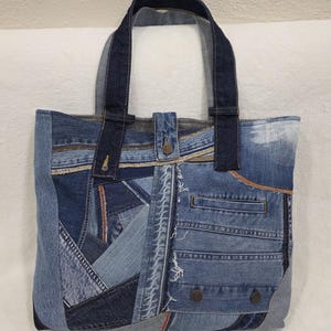 May include: A patchwork denim tote bag. The bag is made from various shades of blue denim, with visible seams and pocket details. The handles are a darker shade of blue. The bag has a rectangular shape and two handles.