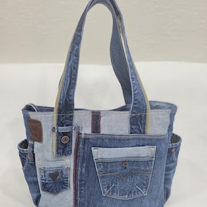 Peut inclure: Un sac fourre-tout en denim fabriqué à partir de jeans recyclés. Le sac comporte plusieurs poches, dont des poches avant et latérales, et un patch en cuir marron. Les anses sont faites du même matériau en denim. Le sac est un mélange de denim bleu clair et foncé.