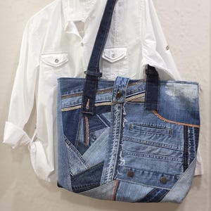 May include: A denim tote bag crafted from recycled jeans. The bag showcases a patchwork design with diverse shades of blue denim, visible pockets, and a dark blue strap. A white shirt hangs in the background.