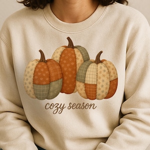 Puede incluir: Sudadera beige con tres calabazas de patchwork en tonos naranja, verde y beige. Las calabazas tienen patrones de estrellas y cuadros. Las palabras "cozy season" están escritas en una fuente cursiva debajo de las calabazas.