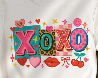May include: White t-shirt featuring a colorful graphic design with the letters "XOXO". The design includes hearts, stars, a smiling face, cherries, and the words "Kiss Me". The letters are in pink, teal, and leopard print.