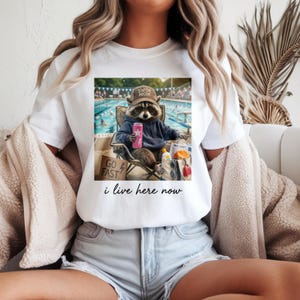 Puede incluir: Camiseta blanca con un estampado de un mapache con una gorra de "Swim Mom" y una sudadera con capucha azul, relajándose junto a una piscina con una lata rosa. El texto "i live here now" está impreso debajo.