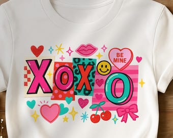 May include: White t-shirt with a colorful graphic design. The design includes the letters "XOXO" in a collage style, along with hearts, lips, a smiley face, cherries, and a bow. The words "BE MINE" are on a heart.