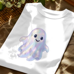 May include: White t-shirt with a cartoon ghost illustration. The ghost is iridescent with a smiling face and dark blue eyes. The design is centered on the shirt, suitable for Halloween or spooky season.