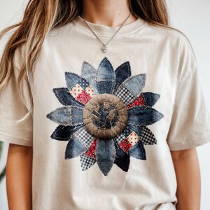 May include: A beige t-shirt featuring a large, denim patchwork sunflower design. The sunflower petals are made of various denim fabrics, including blue, red, and white patterns. The center of the flower is a textured brown fabric.