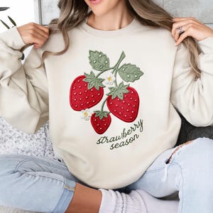 May include: Beige sweatshirt featuring an embroidered strawberry design with three red strawberries, green leaves, and white flowers. The text "strawberry season" is written in a cursive font below the strawberries.