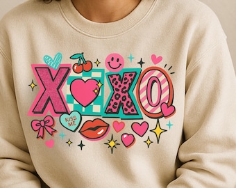 May include: Beige sweatshirt with a colorful graphic design. The design includes the letters "XOXO" in pink, leopard print, and striped patterns, along with hearts, stars, a smiley face, cherries, and the words "Kiss Me".