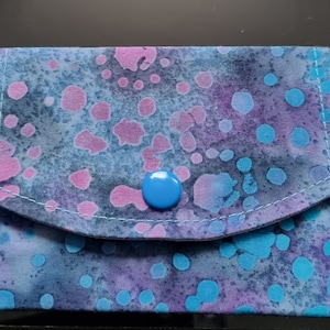 May include: A small blue fabric pouch with a blue button closure. The pouch has a light blue and purple background with white stitching and a scattered pattern of pink and blue circles.