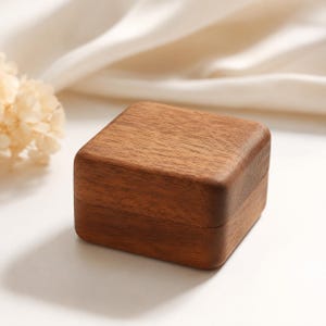 May include: A small, square wooden box with a hinged lid. The box is made of a rich, dark brown wood with a visible grain pattern. The box is sitting on a white surface, with a soft, white fabric and dried flowers in the background.