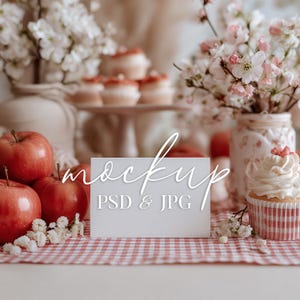 Puede incluir: Una tarjeta de maqueta blanca con la palabra "mockup" en cursiva y el texto "PSD & JPG" debajo. La tarjeta está sobre un mantel a cuadros rojo y blanco, con manzanas rojas, cupcakes y flores en el fondo.