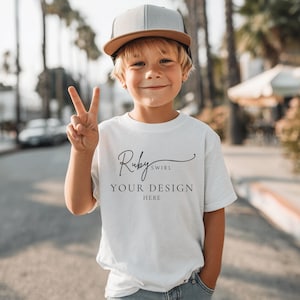 May include: A young child wearing a white t-shirt and a gray and brown baseball cap. The t-shirt has the text "Ruby Swirl YOUR DESIGN HERE" in black script. The child is smiling and holding up two fingers.