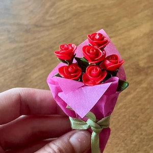 Peut inclure: Un petit bouquet de roses rouges, chacune avec des pétales détaillés, enveloppé dans du papier rose et noué avec un ruban vert. Le bouquet est tenu dans une main. Les roses ont des tiges vertes.
