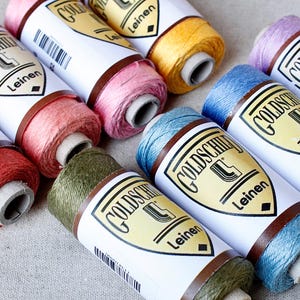 Pode incluir: Uma coleção de bobinas coloridas de linha de linho. As bobinas, rotuladas como "Goldschild Leinen", são em tons de vermelho, rosa, amarelo, verde, azul e roxo. As bobinas estão dispostas sobre uma superfície de cor neutra.