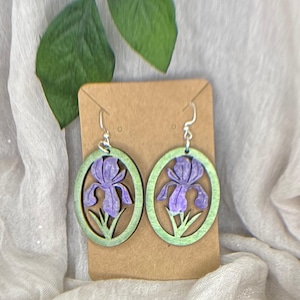 May include: A pair of wooden earrings featuring a purple iris flower design within a green oval frame. The earrings hang from silver-colored hooks and are displayed on a brown card. The background includes a white fabric and green leaves.