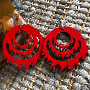 May include: A pair of bright red wooden earrings. Each earring is a circular shape with a jagged, stylized design cut out of the wood. The earrings have silver-colored hooks.