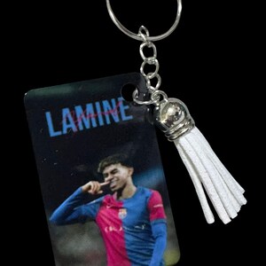 Könnte beinhalten: Ein rechteckiger Schlüsselanhänger mit dem aufgedruckten Bild eines Fußballspielers in einem blau-roten Trikot und dem Namen LAMINE in blauer Schrift darüber. Der Anhänger hat einen silbernen Metallring und eine weiße Kunstlederquaste mit Silberkappe.