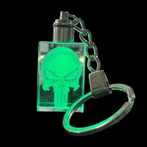 May include: A rectangular clear acrylic keychain featuring an internal laser-etched skull design that glows with a bright green light. The keychain includes a silver-toned metal cap, a short chain, and a circular split ring for attachment.