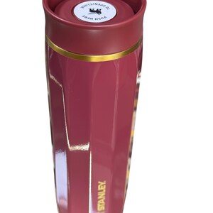 May include: A maroon Stanley travel mug with a gold band. The lid is a darker maroon with the text "TO OPEN/CLOSE" and "PUSH HERE". The mug is cylindrical and has the Stanley logo on the side.