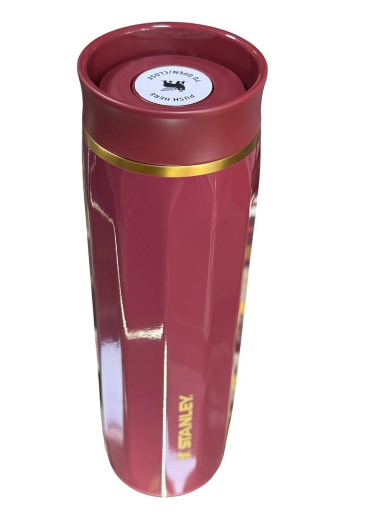 May include: A maroon Stanley travel mug with a gold band. The lid is a darker maroon with the text "TO OPEN/CLOSE" and "PUSH HERE". The mug is cylindrical and has the Stanley logo on the side.