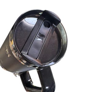 May include: A black Stanley travel mug with a handle and a black lid. The lid has a flip-top closure and a small logo. The mug is made of stainless steel and has a shiny finish.