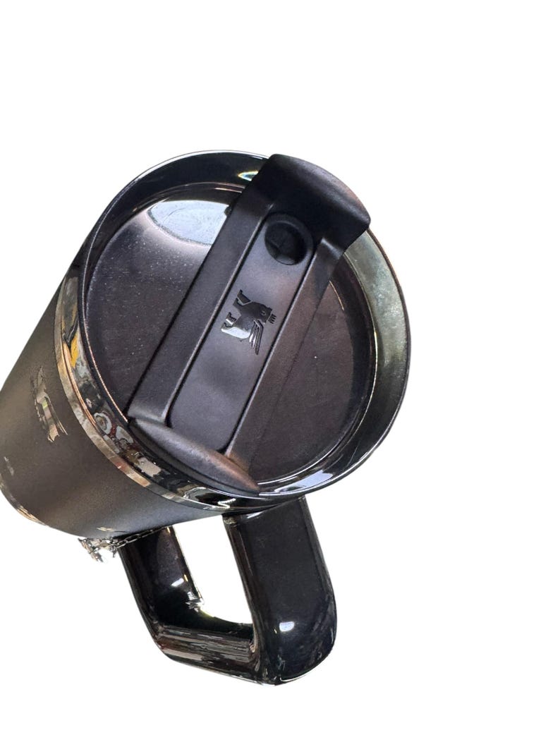 May include: A black Stanley travel mug with a handle and a black lid. The lid has a flip-top closure and a small logo. The mug is made of stainless steel and has a shiny finish.